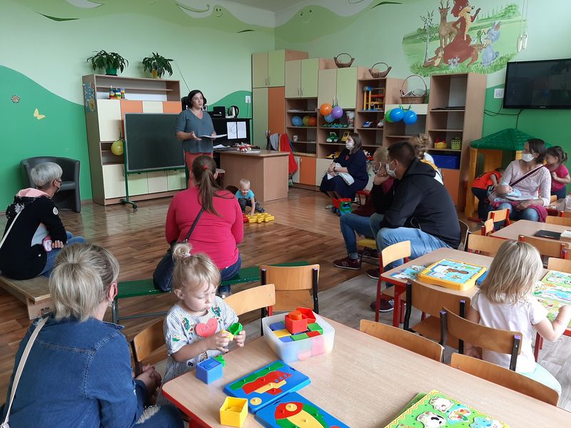 Na pierwszym planie zdjecia widać salę przedszkolną i siedzące w niej dzieci, które układają układanki i puzzle. W tle siedzą rodzice, przed którymi stoi nauczycielka.