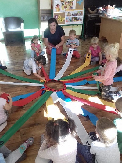 Zdjęcie przedstawia dzieci bawiące się wiatrakiem matematycznym. Każde dziecko trzyma w dłoni kolorową wstążkę. Dzieci wraz z nauczycielką rolują wsążki. 