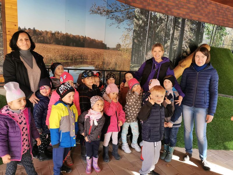 Zdjęcie przedstawia przedszkolaki stojące w grupie. Przy dzieciach stoją dwie nauczycielki i opiekunka. W tle widać ścianę z afiszem przedstawiającym krajobraz Puszczy Białowieskiej.