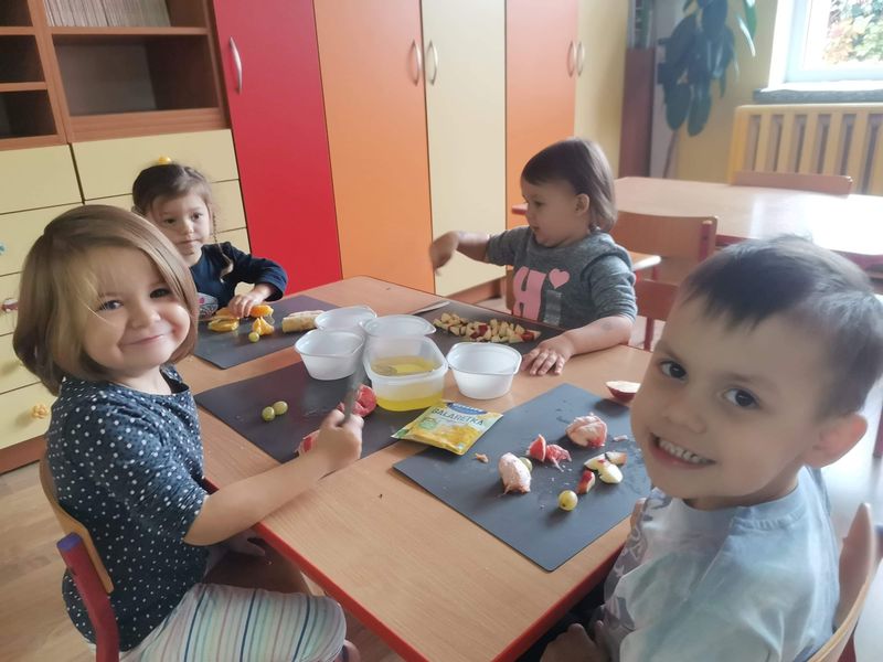 Zdjęcie przedstawia chłopca i trzy dziewczynki siedzące przy stole w przedszkolnej sali. Dzieci mają przed sobą podkładki z własnoręcznie pokrojonymi owocami takimi jak czerwony grejpfrut, pomarańcza, jabłko, winogrono.