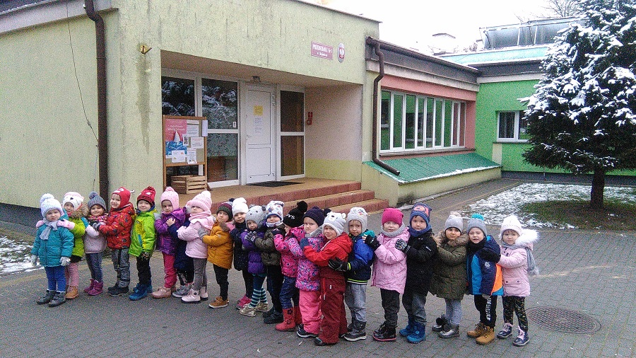 Dzieci z grupy Wróbelki stoją gęsiego przed budynkiem przedszkola.