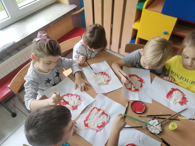 Zdjęcie przestawia pięcioro dzieci, które malują herb Polski biało-czerwonymi farbami. Jest on zrobiony z masy solnej. Dzieci, które są na zdjęciu to zuchy, które należą do 30 Gromady Zuchowej „Leśne Skrzaty”.