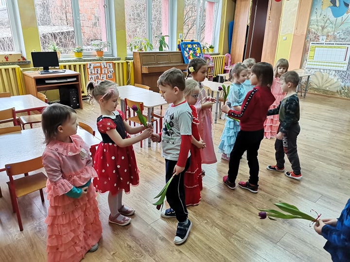 Zdjęcie przedstawia Dzień Dziewczynek w przedszkolu. Chłopcy wręczają różowe tulipany dziewczynkom ubranym w sukienki balowe. 