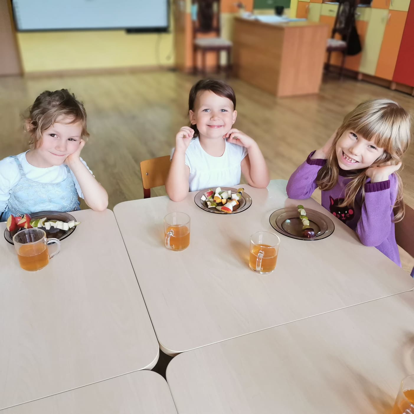 Zdjęcie przedstawia uśmiechnięte dziewczynki siedzące przy stoliku przed którymi leżą szaszłyki z jesiennych owoców ( jabłka, gruszki, śliwki).