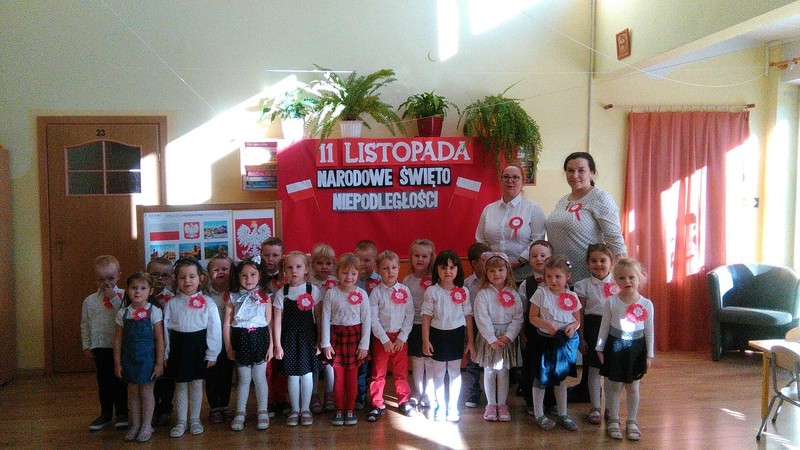 Zdjęcie przestawia grupę dzieci wraz z panią dyrektor i nauczycielką na tle polskiej flagi.