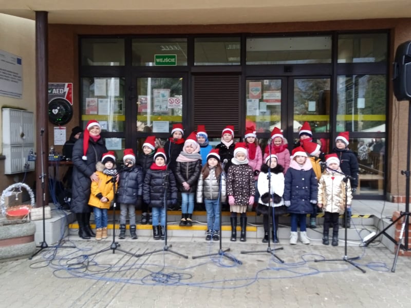 Grupa dzieci przed Urzędem miasta w czerwonych czapkach św. Mikołaja