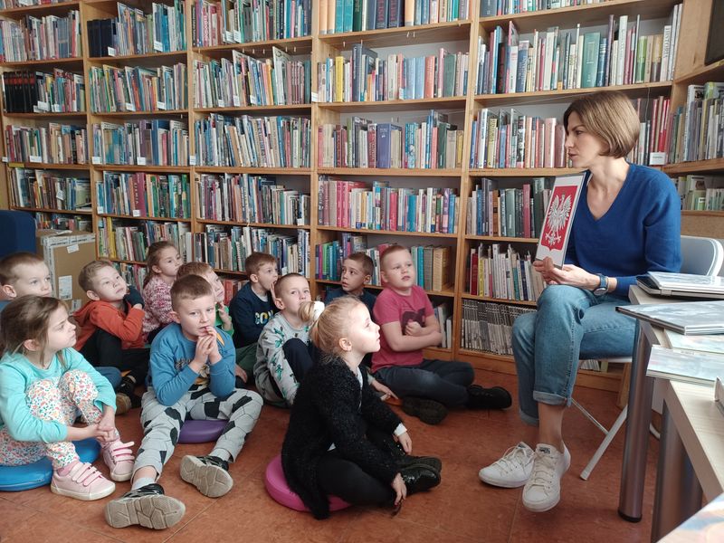 Dzieci siedzą w bibliotece i słuchają bibliotekarki