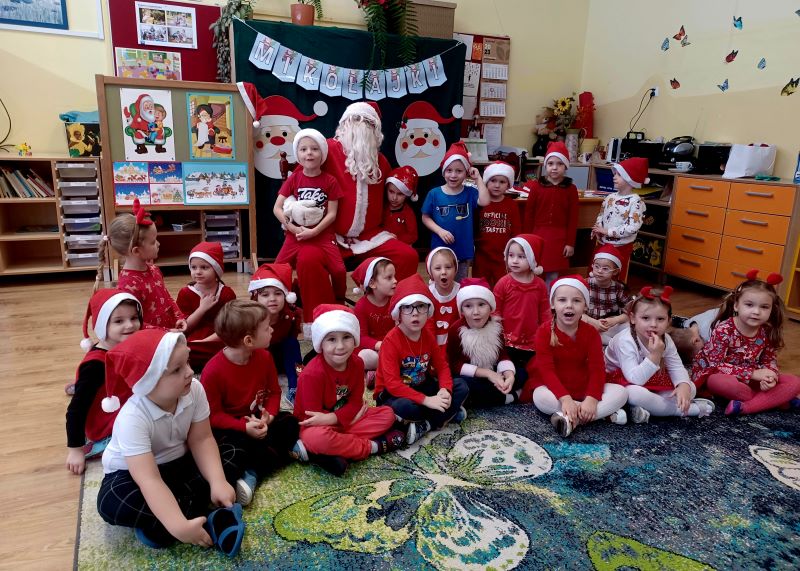 Dzieci ubrane w czerwone świąteczne stroje pozują do zdjęcia. Po środku na krześle siedzi Mikołaj z białą brodą. W tle znajduje się dekoracja z napisem: mikołajki.