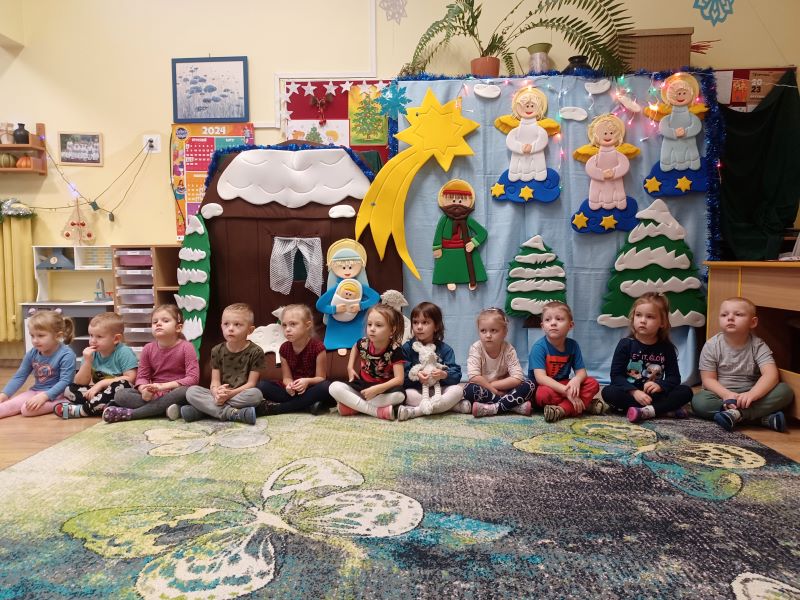 Na zdjęciu przedszkolaki siedzą w szeregu na dywanie w przedszkolnej sali. W tle dekoracja bożonarodzeniowa. 