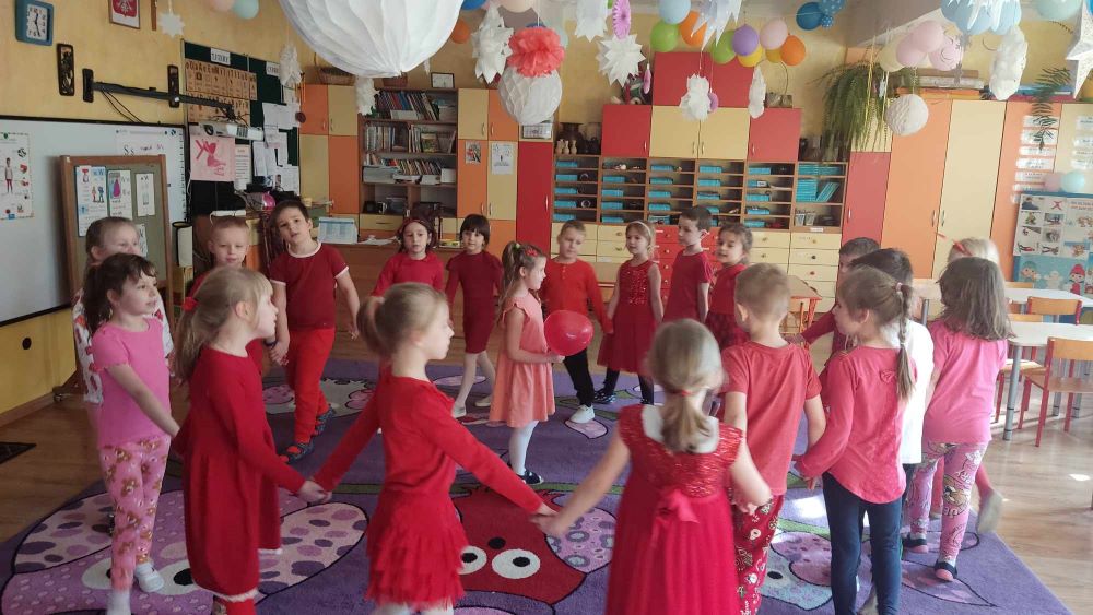 Dzieci trzymając się za ręce tworzą koło, na środku stoi dziewczynka z czerwonym balonem