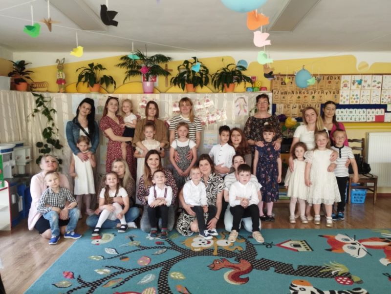 Dzieci i mamy stoją na tle dekoracji, napisu 26 MAJA DZIEŃ MAMY i portretów mam.
