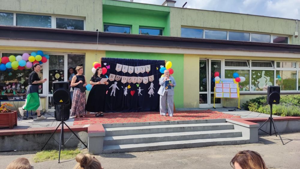 Na przedszkolnym tarasie stoją organizatorki festynu rodzinnego. W tle dekoracja z napisem, kolorowe balony, głośniki.